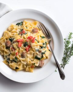 White plate with creamy Tuscan sausage pasta. The bowtie pasta is coated in a creamy sauce, with sausage, spinach, and red peppers adding texture and flavor.