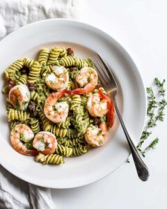 White bowl with Shrimp Pesto Pasta Salad. The pasta is coated in vibrant pesto sauce, topped with shrimp, cherry tomatoes, and crumbled feta cheese.